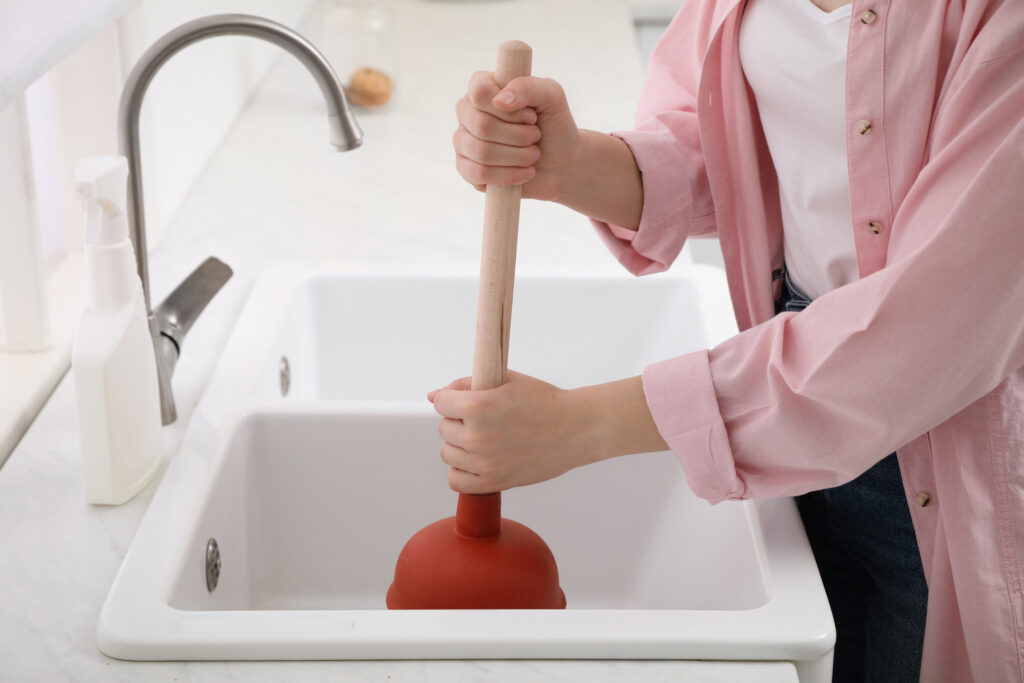 Woman using plunger to unclog sink drain in kitchen, in white porcelain double sink.